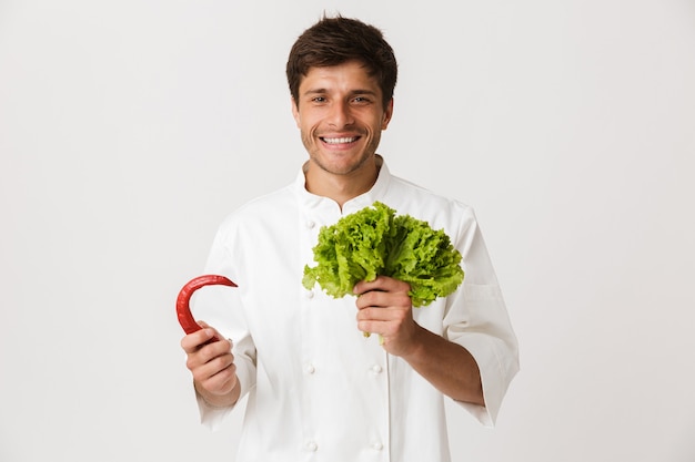 Emotionaler junger Koch lokalisiert auf Weiß, das Salat und Paprikapfeffer hält.