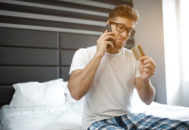 Emotionaler junger Mann am frühen Morgen auf dem Bett. Er telefoniert und schaut auf die Kreditkarte in der Hand. Kerl tragen Pyjama. Tageslicht.