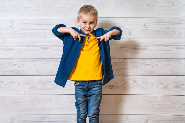 Emotionaler kleiner Junge in blauem Hemd und Jeans, der auf dem Hintergrund der hölzernen Studiowand steht Fröhliches blondes Kind, das seine Finger vor sich zeigt und in die Kamera schaut