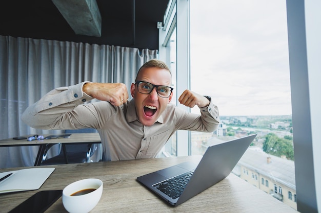 Emotionaler Mann, der am Arbeitsplatz auf die Kamera blickt Ein Mann mit Brille sitzt an einem Desktop mit einem Computer Ein Student sucht nach Inspiration für neue Ideen in einem Bürocafé