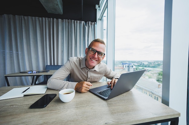 Emotionaler Mann, der am Arbeitsplatz auf die Kamera blickt Ein Mann mit Brille sitzt an einem Desktop mit einem Computer Ein Student sucht nach Inspiration für neue Ideen in einem Bürocafé