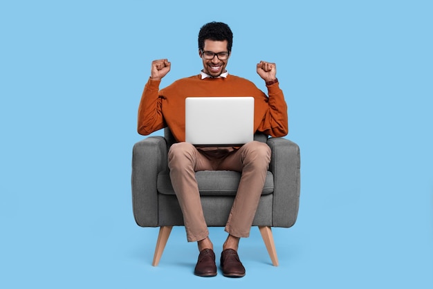 Emotionaler Mann mit Laptop sitzt auf einem Sessel auf hellblauem Hintergrund