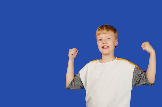 Foto emotionaler neunjähriger teenager zeigt gewinner mit erhobenen händen, isoliert vor blauem hintergrund. kaukasischer junge des porträts im weißen t-shirt. guy hob die hände zu ehren des sieges. platz kopieren