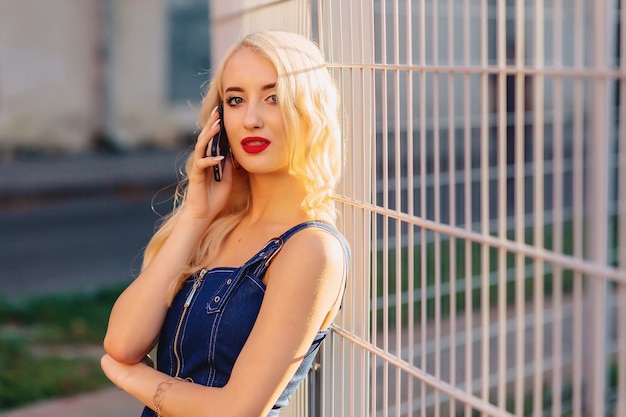 Emotionales blondes attraktives Mädchen in der Sonnenbrille mit Telefon im Sommersonnenschein städtisch
