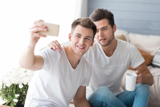 Emotionales junges homosexuelles Paar, das lächelt und Selfie nimmt, während es auf dem Sofa sitzt.