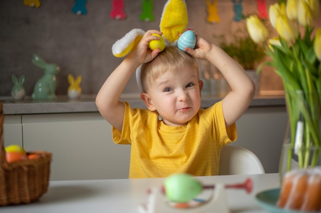 Emotionales Porträt eines fröhlichen kleinen Jungen mit Hasenohren am Ostertag, der fröhlich lacht und mit bunten Ostereiern spielt, die an einem Tisch in der Küche sitzen