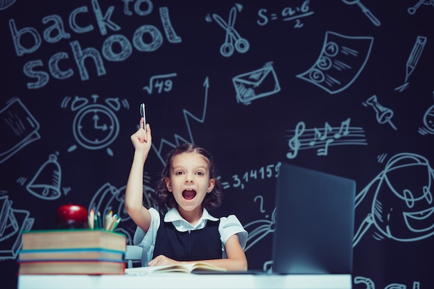 Emotionales Schulmädchen hebt die Hand mit dem Stift, der mit dem Laptop-Fernunterricht sitzt