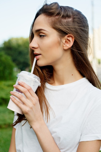 Emotionen, Lifestyle, Schönheit, Menschen, Food-Konzept-Urban Portrait einer jungen attraktiven brünetten Frau in einem weißen T-Shirt, die bei Sonnenuntergang Milchshake durch einen Röhrensommer trinkt