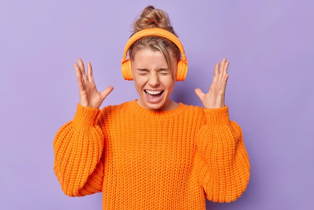 Emotiva mujer loca exclama en voz alta mantiene las manos levantadas se siente enojada escucha música con sonido fuerte usa audífonos inalámbricos en las orejas para cancelación de ruido vestida con suéter de punto posa en el interior