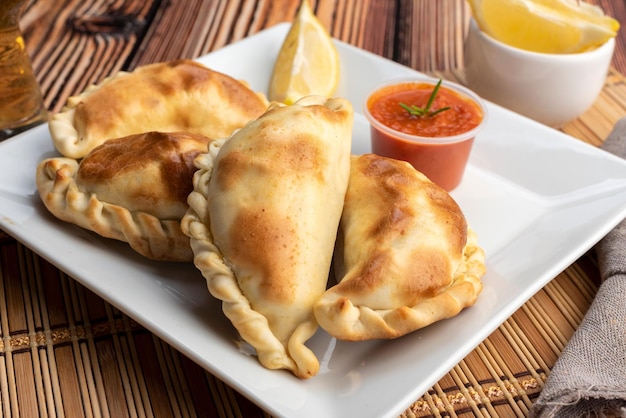 Empanadas caseras argentinas sobre plato cuadrado blanco, salsa roja y rodajas de limón sobre mesa de madera