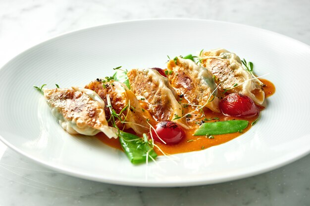 Empanadillas fritas o Gyoza japonesa en salsa amarilla, tomates cherry y guisantes, servidas en un plato blanco sobre una mesa de mármol. Cocina panasiática. Comida de restaurante