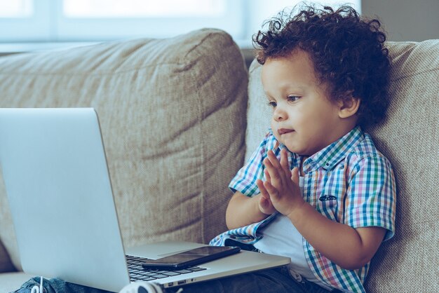 ¡Empecemos a trabajar! Pequeño bebé africano mirando su computadora portátil mientras está sentado en el sofá en casa