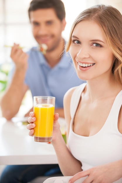 Empezando por la mañana juntos. Apuesto joven sosteniendo un vaso de jugo de naranja y mirando por encima del hombro mientras desayuna con su novia