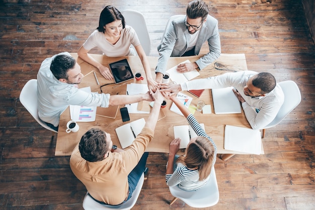 Empezar una nueva jornada laboral en equipo. Vista superior del grupo de seis jóvenes tomados de la mano juntos y sonríen mientras están sentados en el escritorio de la oficina