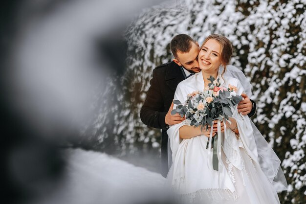 Foto empfindliches porträt eines glücklichen jungvermählten der bräutigam umarmt die braut im winterpark lächelnde braut in hochzeitskleidung und weißem poncho der brüder ist in einem schwarzen mantel gekleidet weitwinkel freiraum