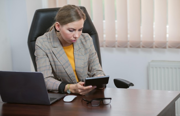Empfindungsfähige Chefin berechnet die Kosten auf einem Taschenrechner, während sie in ihrem Büro am Tisch sitzt