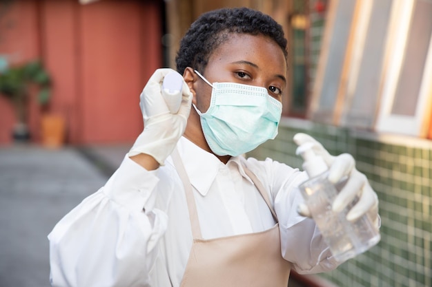 Empleada africana negra o dueña de una tienda con dispensador de gel de alcohol, desinfectante de manos y pistola de escáner térmico para verificar la temperatura, nuevo concepto de práctica comercial normal