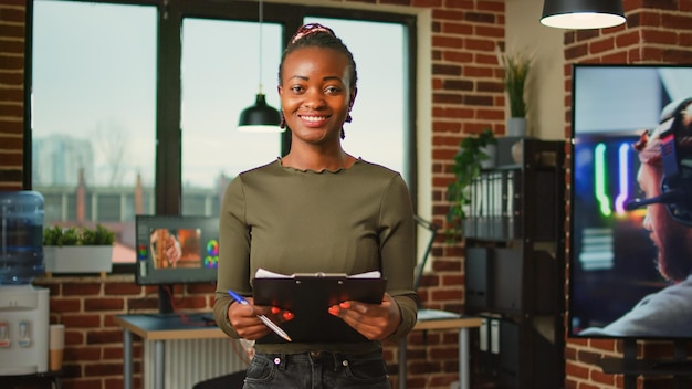 Empleada afroamericana leyendo trabajo de documentación en la oficina de la agencia digital, diseñadora de arte sonriente. Mujer mirando los archivos del portapapeles antes de usar la interfaz de software 3d.