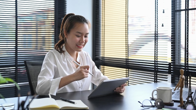 Empleada asiática sentada en su lugar de trabajo y usando una tableta digital revisando datos financieros