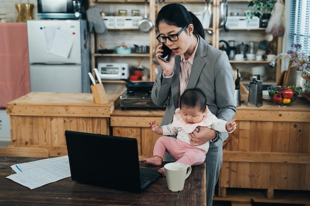 una empleada coreana con un niño está respondiendo llamadas y mirando información en una computadora portátil. una mujer de carrera asiática de pie se lastimó la mano y puso al bebé en la mesa mientras teletrabajaba con teléfono y computadora.