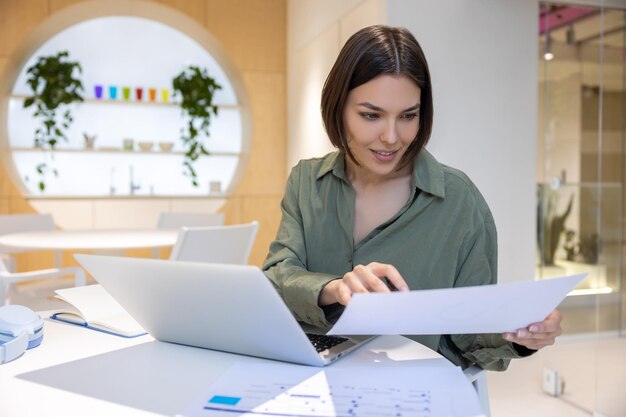 Foto empleada corporativa enfocada sentada en la computadora portátil mirando un documento en sus manos