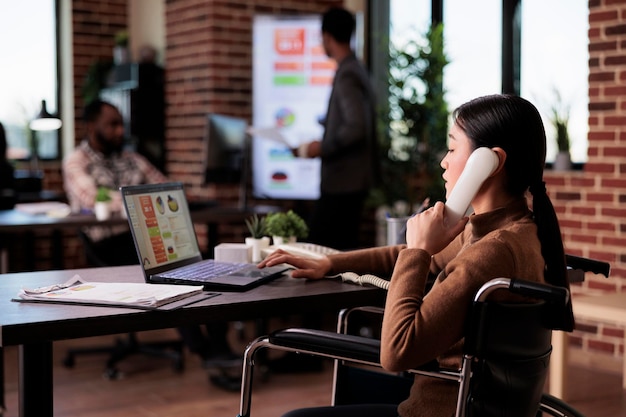 Empleada con problemas de salud que responde a una llamada de teléfono fijo en una oficina adaptada para discapacitados. Uso del teléfono para conversaciones remotas, con discapacidad crónica en silla de ruedas.