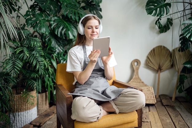 Una empleada del taller de flores planta para el hogar usando una tableta y auriculares sentados