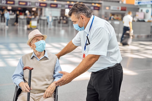 Empleado del aeropuerto hablando con un turista senior con un bastón sentado en una silla de ruedas