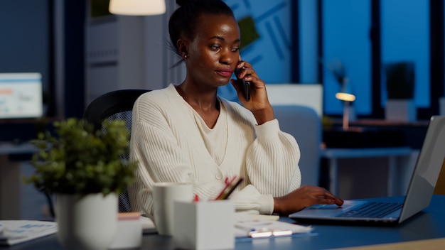 Empleado africano hablando por teléfono mientras trabaja en la computadora portátil a altas horas de la noche