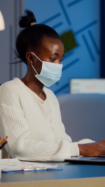 Empleado africano con mascarilla de protección trabajando en horas extraordinarias de portátil