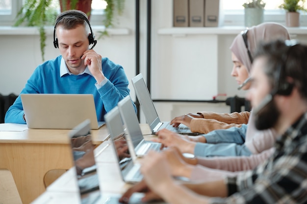 Empleado del centro de contacto joven serio que ajusta el micrófono en los auriculares mientras trabaja con las quejas de los clientes