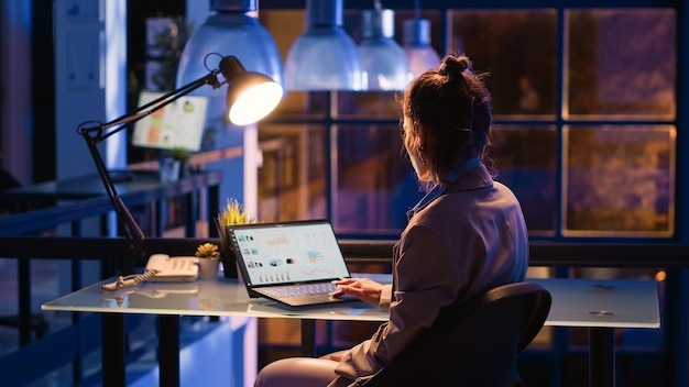 Empleado corporativo que trabaja horas extras en el informe de gráficos en una computadora portátil, planificando la inversión de la empresa por la noche en la oficina. Gerente femenina siguiendo análisis para aumentar las ganancias. Disparo de trípode.