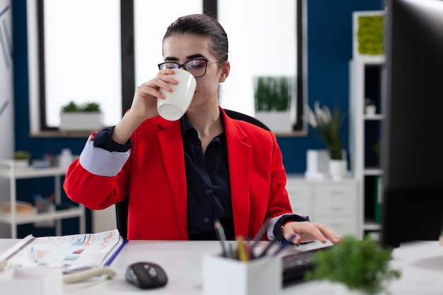 Empleado con gafas en el escritorio de inicio bebiendo de una taza blanca. Mujer de negocios con chaqueta roja tomando café o té. Propietario de una pequeña empresa haciendo una pausa en el trabajo con la computadora.