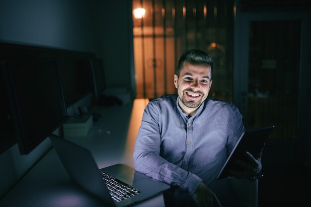 Empleado con gran sonrisa en su rostro usando tableta mientras está sentado en la oficina a altas horas de la noche.