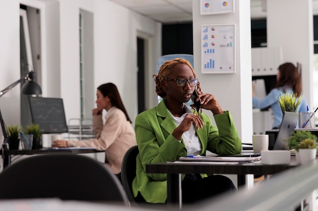 Empleado hablando por teléfono inteligente en un espacio abierto de coworking, haciendo una llamada telefónica con socios comerciales. Joven afroamericana sentada en el escritorio del lugar de trabajo, oficinistas ocupados