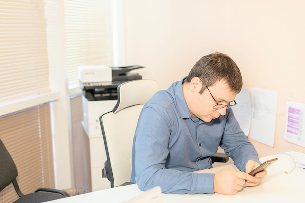 Empleado de hombre con camisa azul y anteojos sentado en el escritorio de la oficina con teléfono inteligente usando aplicaciones de tecnología digital para el desarrollo empresarial Concepto de día de trabajo ocupado de negocios