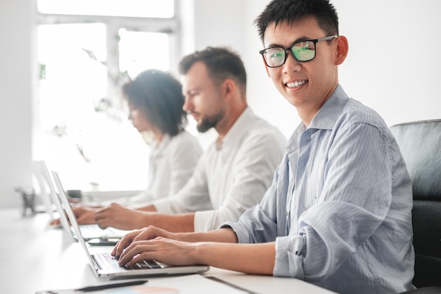 Empleado masculino asiático sonriente que trabaja en una computadora portátil cerca de diversos colegas en la oficina