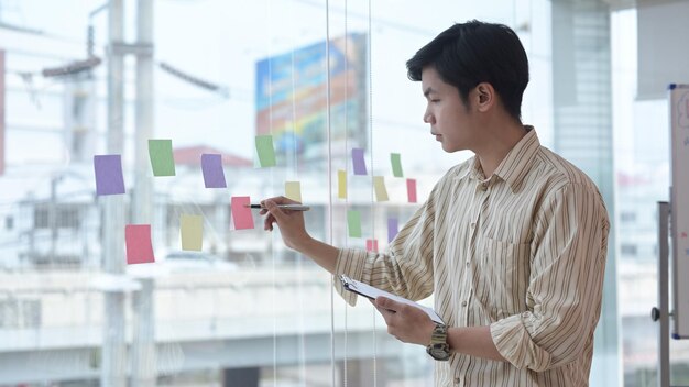 Empleado masculino pensativo escribiendo información sobre notas adhesivas coloridas para desarrollar un proyecto empresarial en la oficina