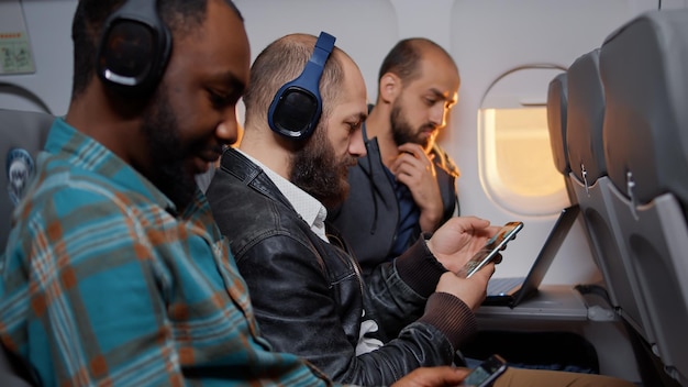 Empleado masculino sentado en un vuelo de avión durante la puesta de sol, navegando por Internet en un smartphone en un viaje de trabajo en avión. Trabajando en el teléfono móvil y volando con una aerolínea comercial. Disparo de mano.