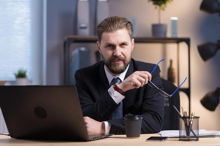 Empleado de oficina barbudo de cabello castaño posando junto a una computadora portátil
