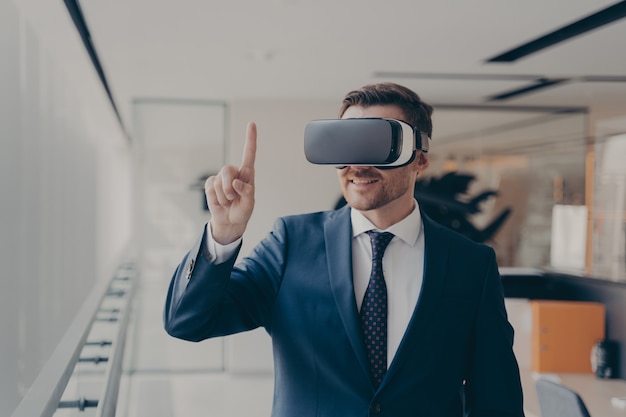 Empleado de oficina barbudo sonriente en traje con gafas VR trabajando en realidad aumentada en la oficina, empresario en gafas 3d interactuando con el ciberespacio en el trabajo, señalando con el dedo