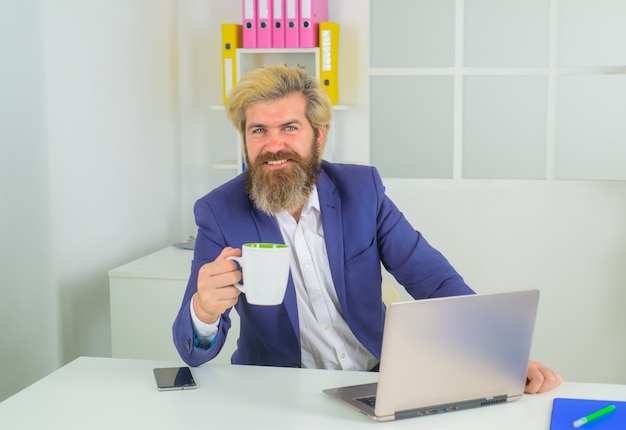 Empleado de oficina. Empresario barbudo con café en la oficina. Hombre de negocios en la computadora portátil. Hombre de negocios con ordenador portátil. Liderazgo. CEO. Experto en negocios.