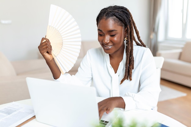 Empleado de oficina que sufre de calor aire caliente y sofocante trabajando en una computadora portátil desde casa sin acondicionador en el día de verano Mujer de negocios africana enfriándose con un ventilador de mano en el lugar de trabajo