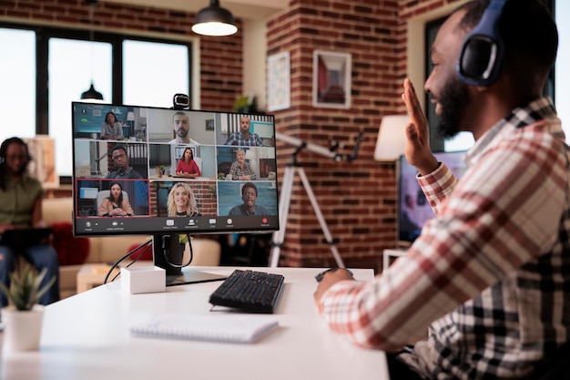 Foto empleado que trabaja a distancia saludando al equipo en videoconferencia en una computadora personal en la sala de estar. hombre afroamericano en una llamada por internet saludando a sus colegas mientras su novia está trabajando en el sofá.