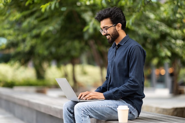 Empleado remoto de hombre oriental positivo trabajando en el parque usando una computadora portátil