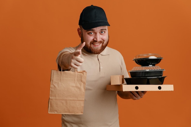 Foto empleado repartidor con gorra negra y uniforme en blanco sosteniendo recipientes de comida con bolsa de papel mirando a la cámara apuntando con el dedo índice a la cámara sonriendo alegremente