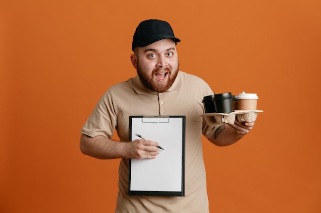 Empleado repartidor con gorra negra y uniforme de camiseta en blanco sosteniendo tazas de café y portapapeles con bolígrafo mirando a la cámara feliz y sorprendido de pie sobre fondo naranja