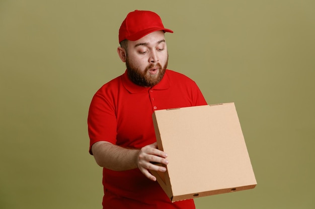 Empleado repartidor con gorra roja camiseta en blanco uniforme sosteniendo caja de pizza abriendo la caja mirando dentro sorprendido de pie sobre fondo verde