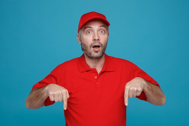 Empleado repartidor con gorra roja uniforme de camiseta en blanco apuntando con los dedos índices hacia abajo mirando a la cámara sorprendido de pie sobre fondo azul