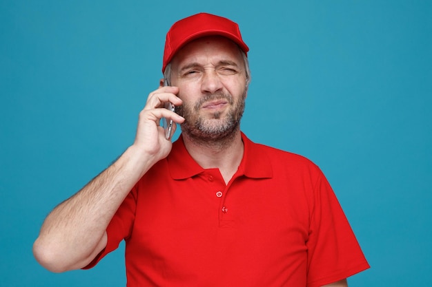 Empleado repartidor con gorra roja uniforme de camiseta en blanco hablando por teléfono móvil con aspecto disgustado haciendo la boca torcida de pie sobre fondo azul